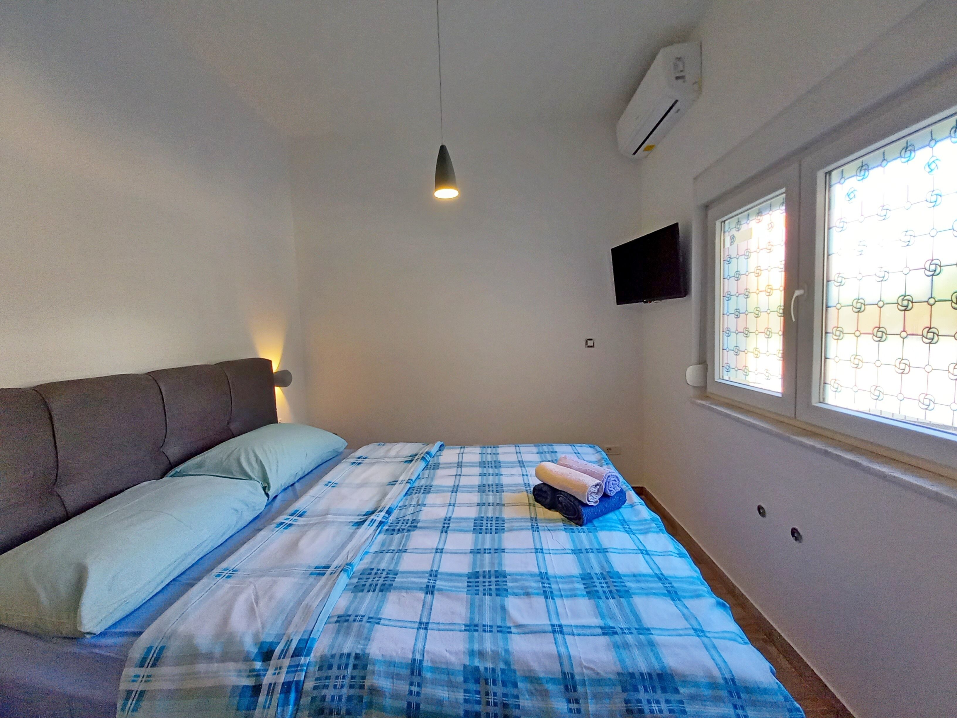 A bedroom with a bed, a bedside lamp, an air conditioner, a TV, and a window with a grid pattern.