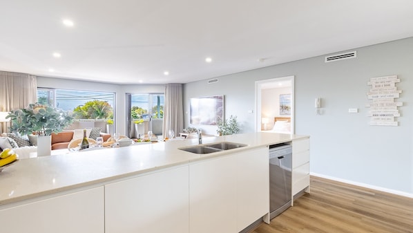 A modern kitchen with a central island, a dining area with a view, and a living space with a sofa and plants.