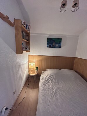 Ein Schlafzimmer mit einem Bett, einem Nachttisch mit Lampe, einer kleinen Ablage mit Büchern und einem Bild an der Wand.