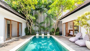 A rectangular swimming pool with a wooden deck surrounding it, flanked by two rooms with sliding glass doors and a small seating area with cushions. - Villa Lapis by Villa Finder (Canggu)
