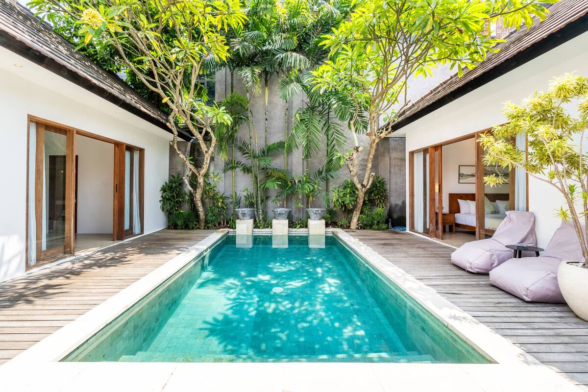 A rectangular swimming pool with a wooden deck surrounding it, flanked by two rooms with sliding glass doors and a small seating area with cushions.