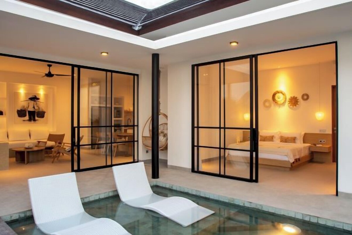 Ein modernes Interieur mit einem gläsernen Schlafzimmer, einem Poolbereich mit Liegestühlen und einem Essbereich mit Deckenventilator.