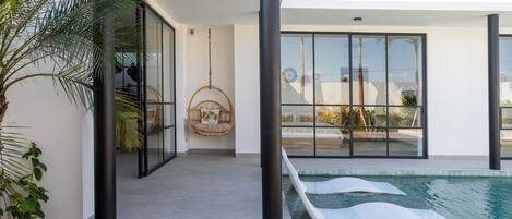Una casa moderna con puerta de vidrio que da a una zona de piscina, una silla colgante y vistas del paisaje circundante.