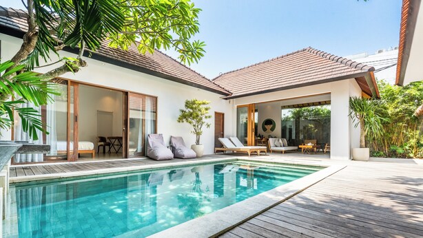 A modern poolside area with a wooden deck, a clear swimming pool, and a covered seating area with lounge chairs and a table. - Villa Lapis by Villa Finder (Canggu)