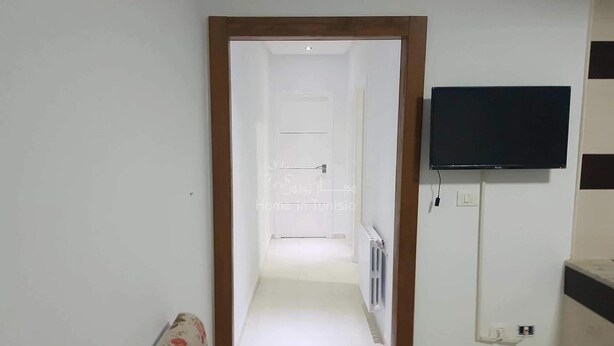 Ein Zimmer mit einer holzrahmenverzierten Tür, die zu einem Flur führt, einem an der Wand befestigten Flachbildfernseher und einem Heizkörper an der angrenzenden Wand.
