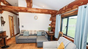 A cozy living room with a grey sofa, a smaller grey armchair, wooden beams on the ceiling, a large window with curtains, and a wooden table with a plant. - Cedar Cabin at Salford Court Farm Fishing Cabins (Worcester)