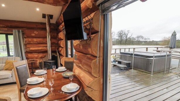 A wooden cabin interior with a dining table set for four, a fireplace, and a view of a wooden deck and outdoor area. - Cedar Cabin at Salford Court Farm Fishing Cabins (Worcester)