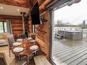 Interior de una cabaña de madera con una mesa de comedor preparada para cuatro personas, una chimenea y vistas a una terraza de madera y zona exterior.