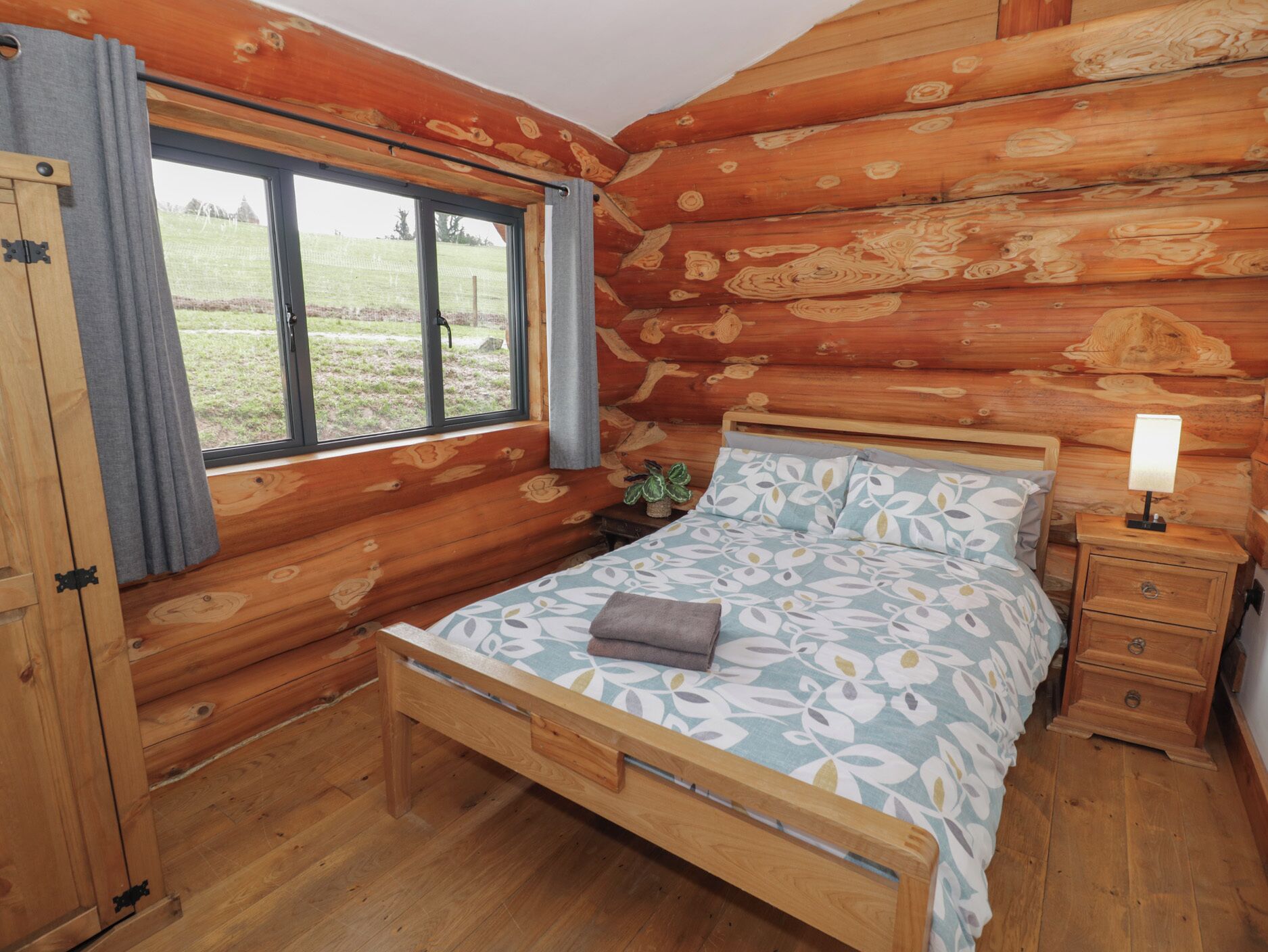 A log cabin bedroom with a bed, bedside table, and a window with curtains.
