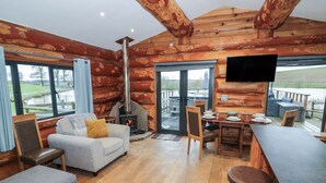 A log cabin interior with a fireplace, seating area, dining table, and large windows offering a view of the outdoors. - Cedar Cabin at Salford Court Farm Fishing Cabins (Worcester)