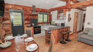 A log cabin-style kitchen with a dining area, wooden beams, a stove, and a view through the window. - Cedar Cabin at Salford Court Farm Fishing Cabins (Worcester)