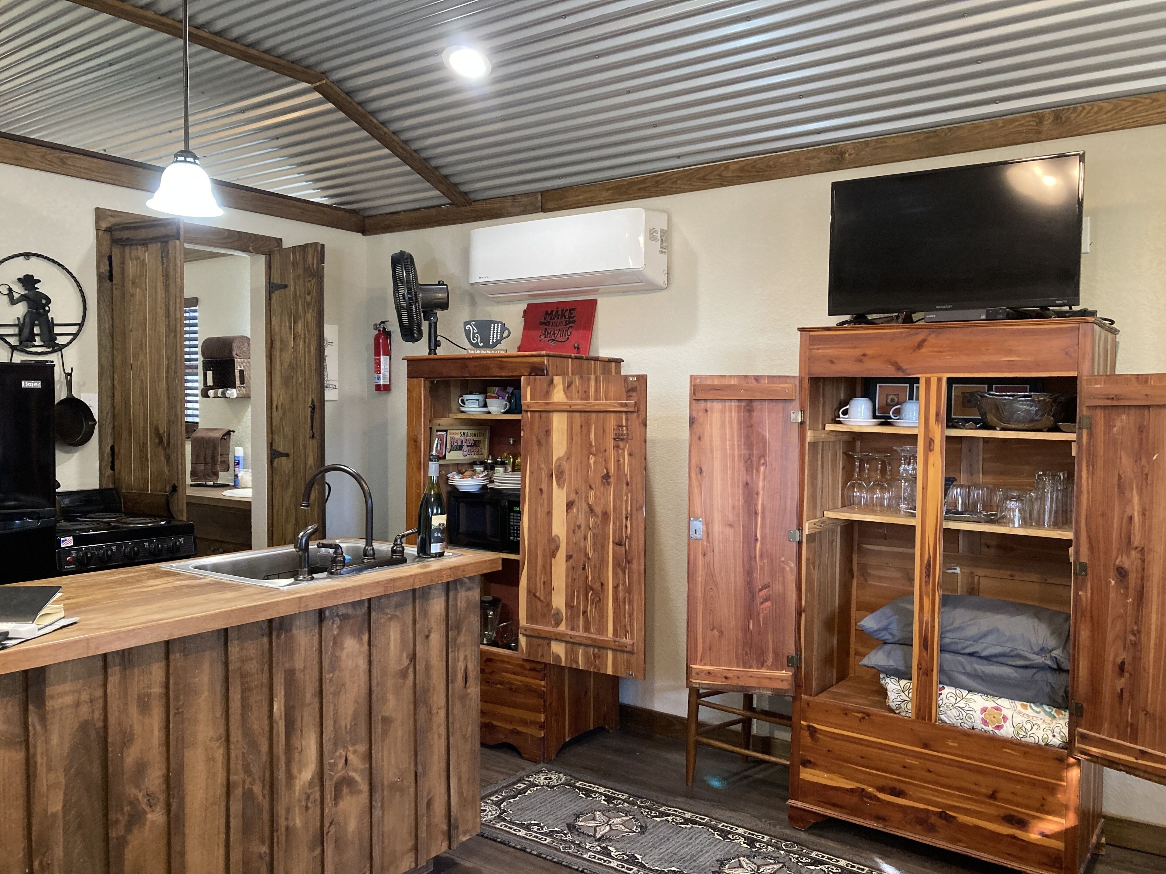 Une cuisine rustique avec des meubles en bois, un poêle et un évier. Un téléviseur est fixé au mur et un ventilateur de plafond est installé.