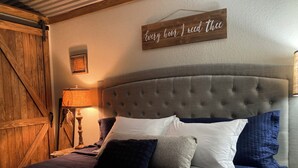A bedroom with a tufted headboard, a bed with blue and white bedding, a wooden wall hanging, and a rustic wooden door. - Sunrise Sunset Peaceful Country Cabin  (Bruceville)