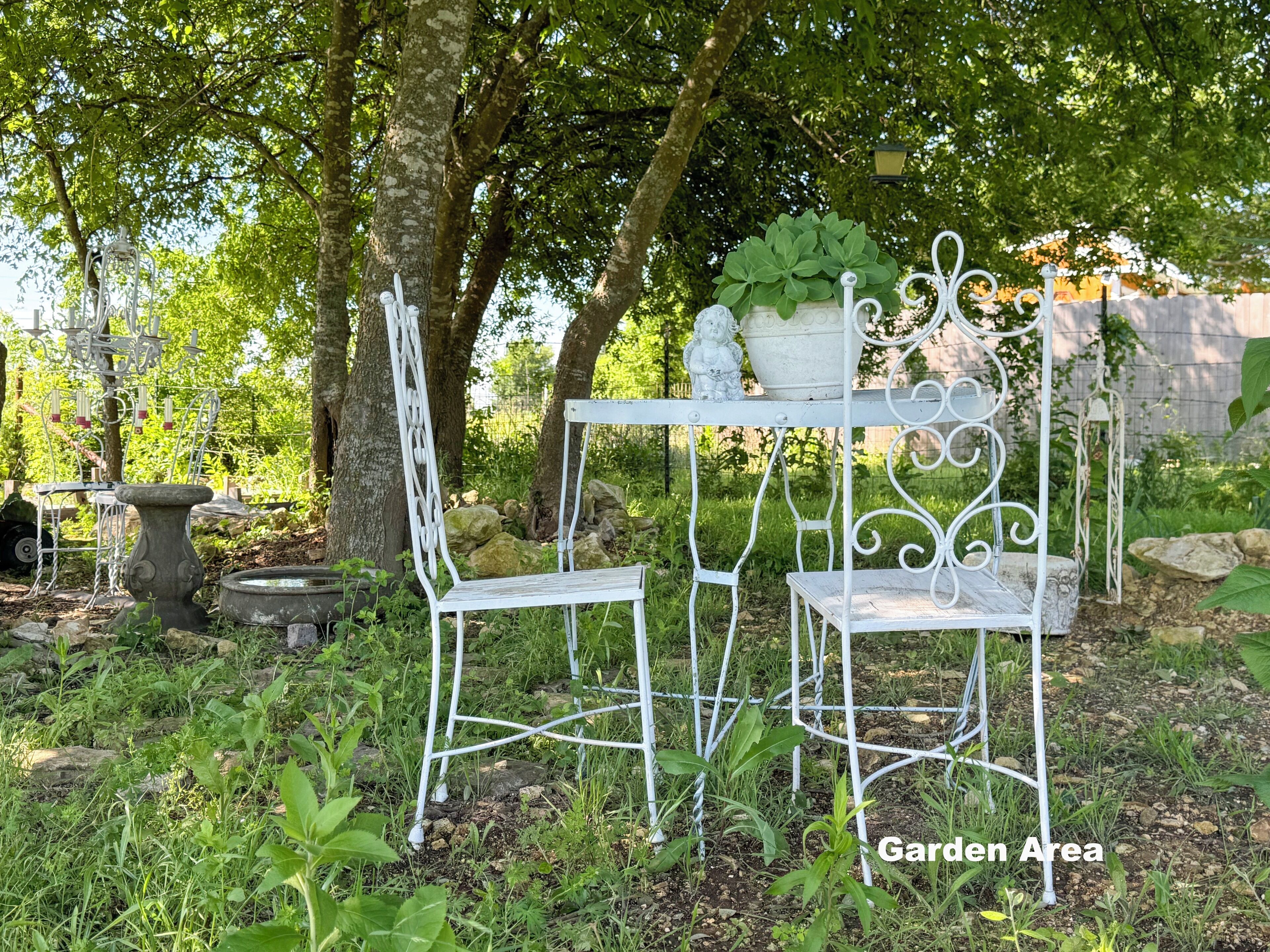 A garden area with a white metal table and chairs, surrounded by greenery and trees.