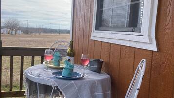 Um deck de madeira com uma mesa posta para dois, uma cadeira e uma janela com vista para uma paisagem rural.