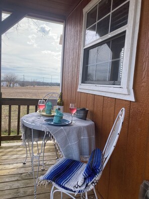 Une terrasse en bois avec une table dressée pour deux personnes, une chaise et une fenêtre donnant sur un paysage rural.