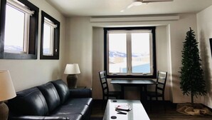 Un salon avec un canapé en cuir noir, une petite table, une table à manger avec des chaises, un ventilateur de plafond et une fenêtre donnant sur un paysage enneigé.