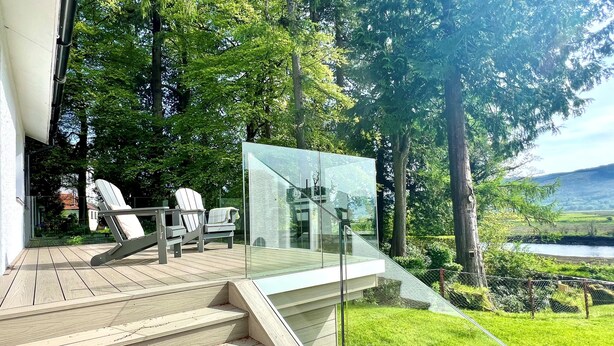 A wooden deck with outdoor seating, a glass balustrade, and a view of a river and greenery.