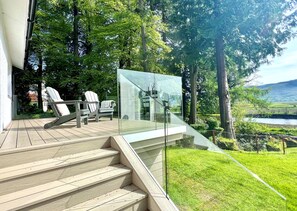 Eine Holzterrasse mit Außenmöbeln, einem Glasgeländer und Blick auf einen Fluss und Grünflächen.