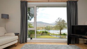 A living room with a sofa, a TV, and a large window offering a view of a lake and hills.