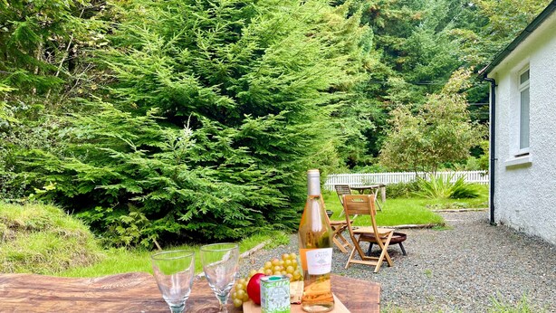 A wooden table outdoors with a bottle of wine, two glasses, and a fruit bowl. - Nursery Cottage - Benmore -  woodland setting, sleeps 4 guests (Benmore, Dunoon)