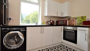 A compact kitchen with a washing machine, a microwave, and a stove. - Nursery Cottage - Benmore -  woodland setting, sleeps 4 guests (Benmore, Dunoon)