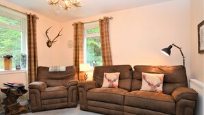 A living room with two brown sofas, a wooden side table, a deer head mounted on the wall, plaid curtains, and a lamp. - Nursery Cottage - Benmore -  woodland setting, sleeps 4 guests (Benmore, Dunoon)
