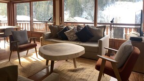 A cozy cabin interior with a large window overlooking a snowy landscape, wooden furniture, and a fireplace.