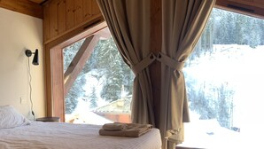 A wooden cabin bedroom with a bed, curtains, and a window overlooking a snowy forest.