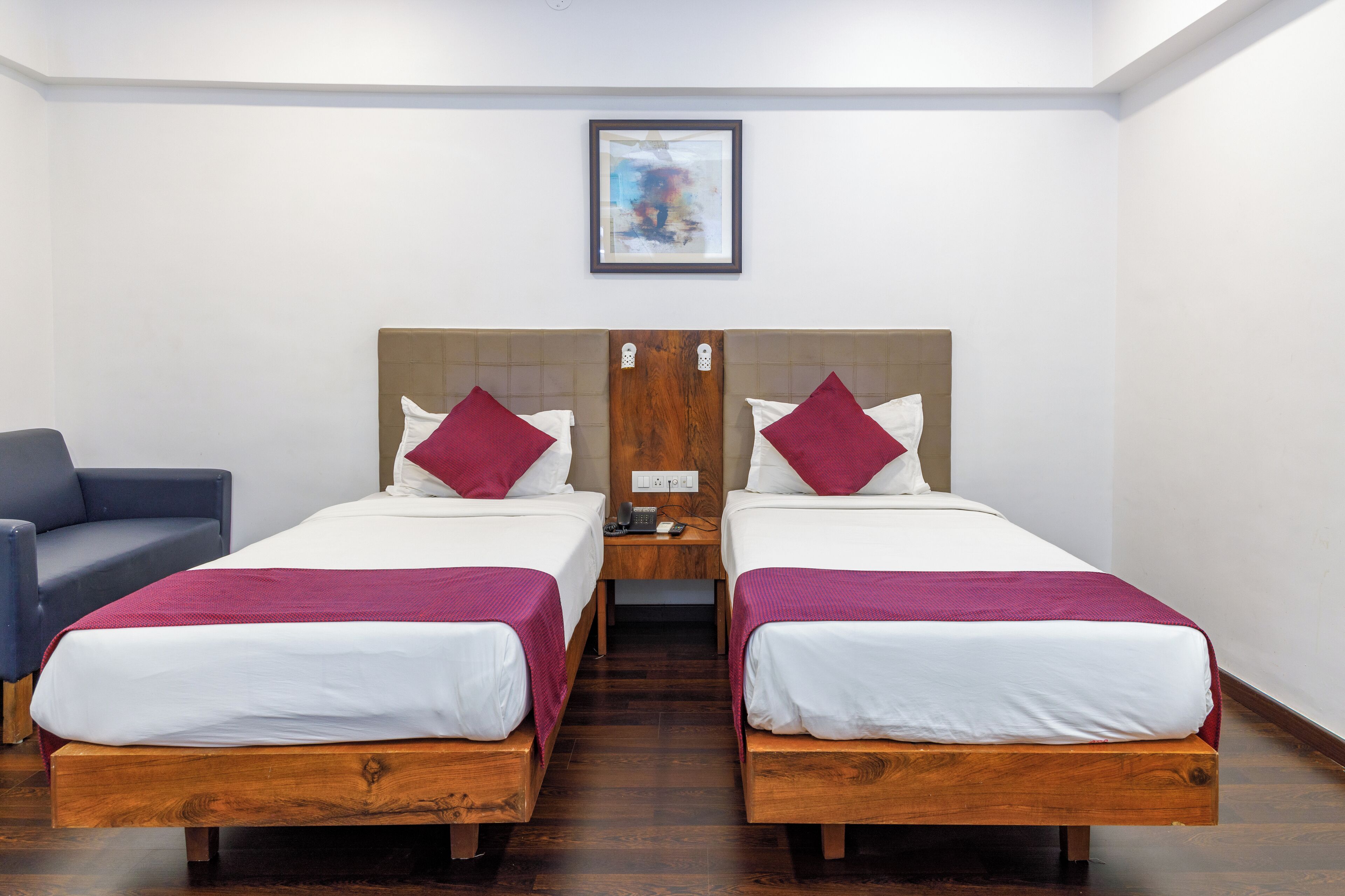 A hotel room with two wooden beds, white bedding, and maroon pillows. There is a wooden nightstand between the beds with a phone and a small table. A blue chair is to the left of the beds.