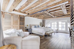 Un dormitorio con techo de madera, cama con ropa de cama blanca, un banco, una mesita de noche y una ventana con vistas.