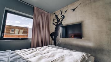 A bedroom with a bed, a window, a curtain, a wall-mounted TV, and a brick wall outside.