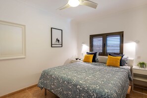 Un dormitorio con cama, dos mesitas de noche, ventilador de techo, ventana con persianas y un cuadro enmarcado en la pared.
