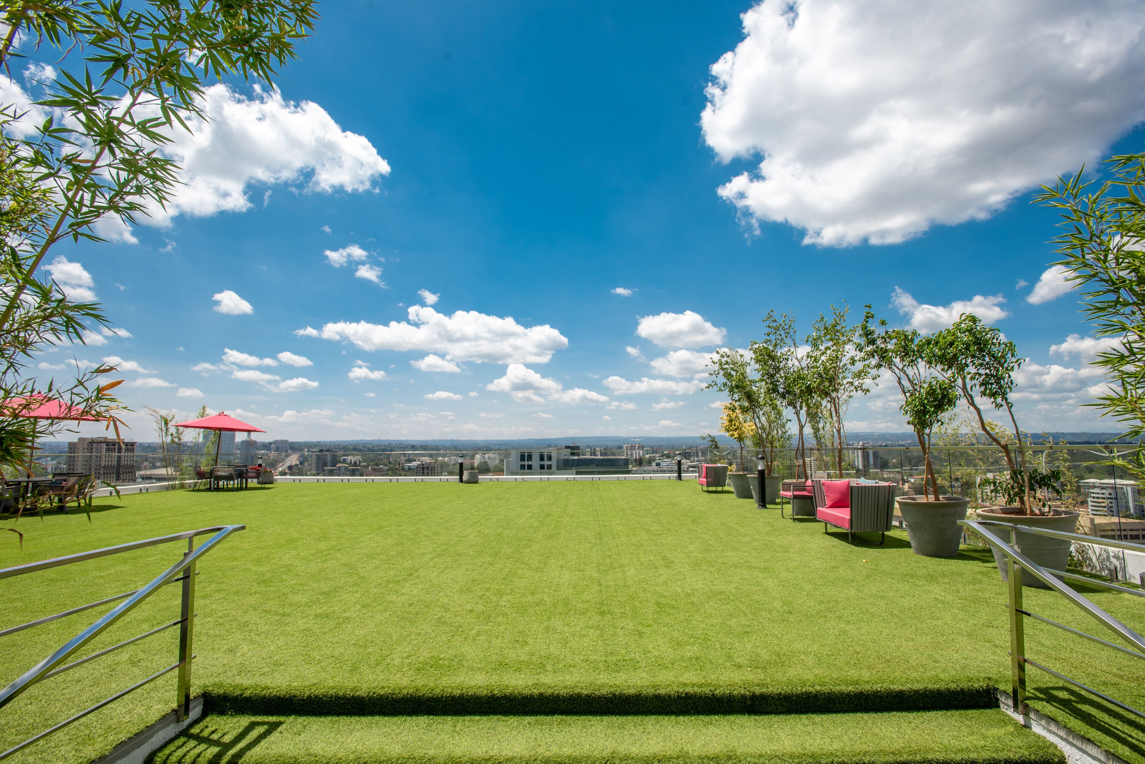 A rooftop terrace with green artificial grass, outdoor furniture, potted plants, and a clear view of the city skyline.