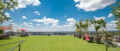 A rooftop terrace with green artificial grass, outdoor furniture, potted plants, and a clear view of the city skyline.