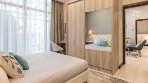 Ein modernes Hotelzimmer mit einem großen Bett, einer Sitzecke mit Sofa und Sesseln, einem Holzschrank und Blick durch ein Fenster mit durchscheinenden Vorhängen.