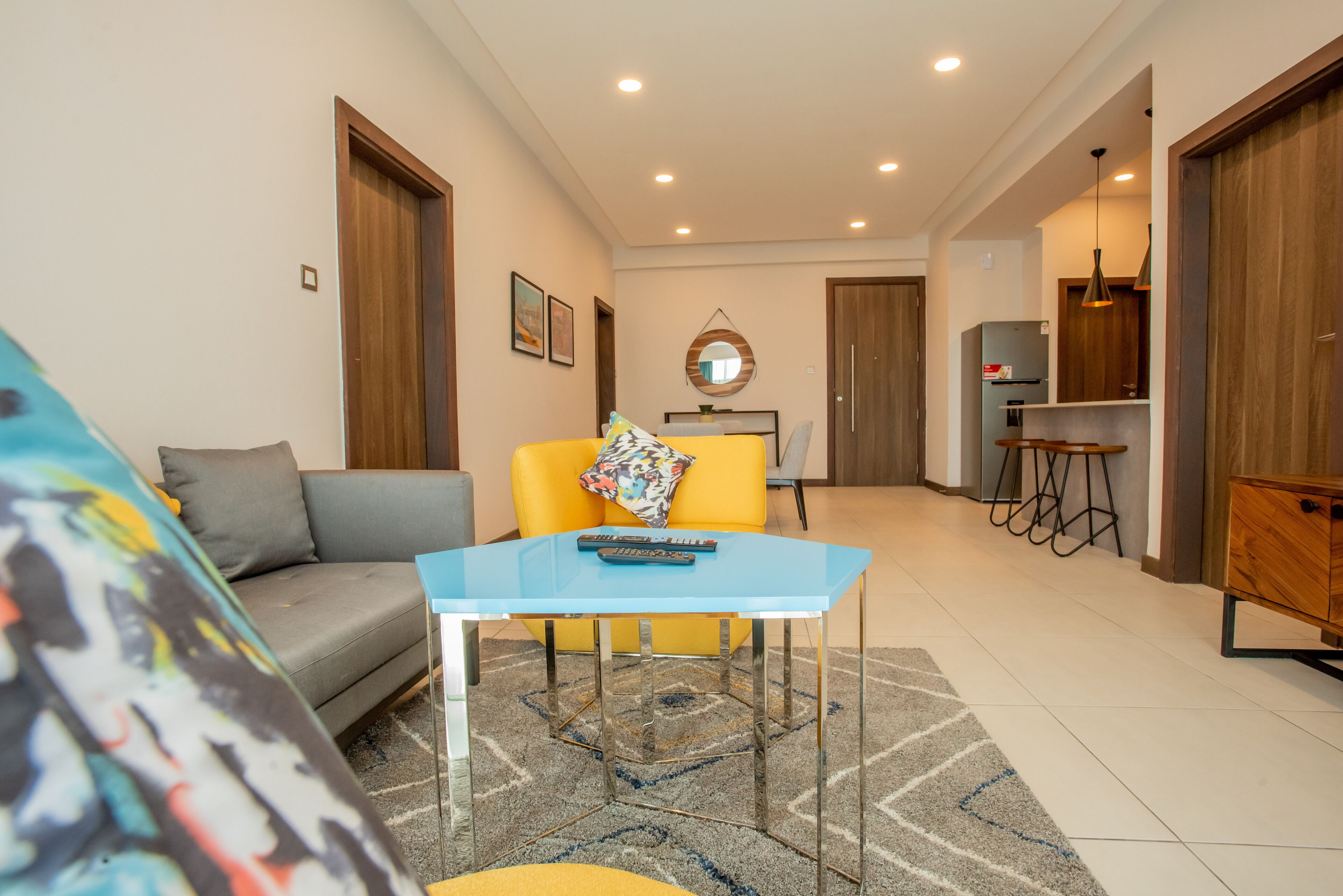 A modern living room with a sofa, a yellow chair, a glass table, and a kitchen area in the background.