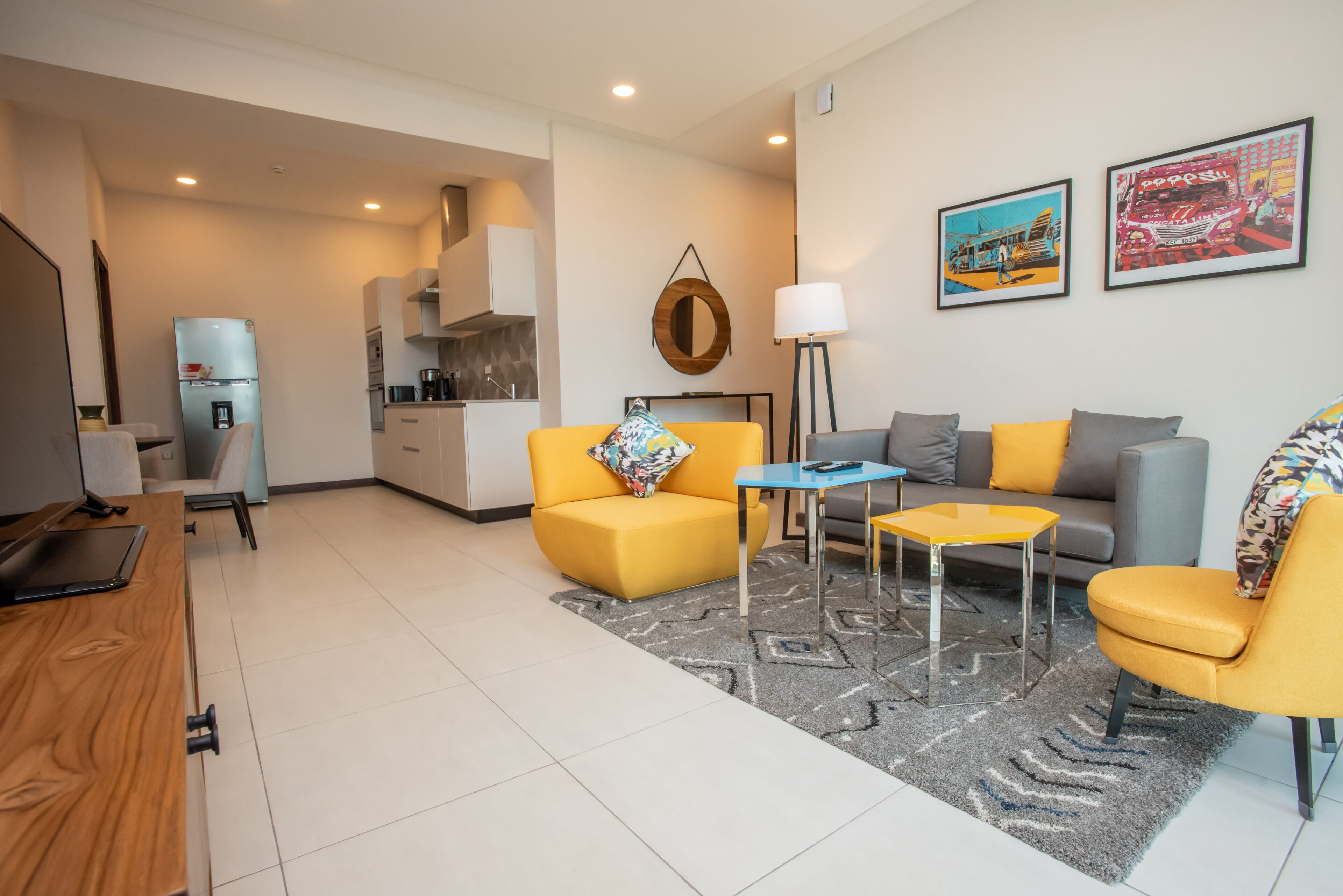 A modern living room with a yellow sofa, a grey sofa, and a blue coffee table. There is a kitchen area in the background with a refrigerator and a sink.