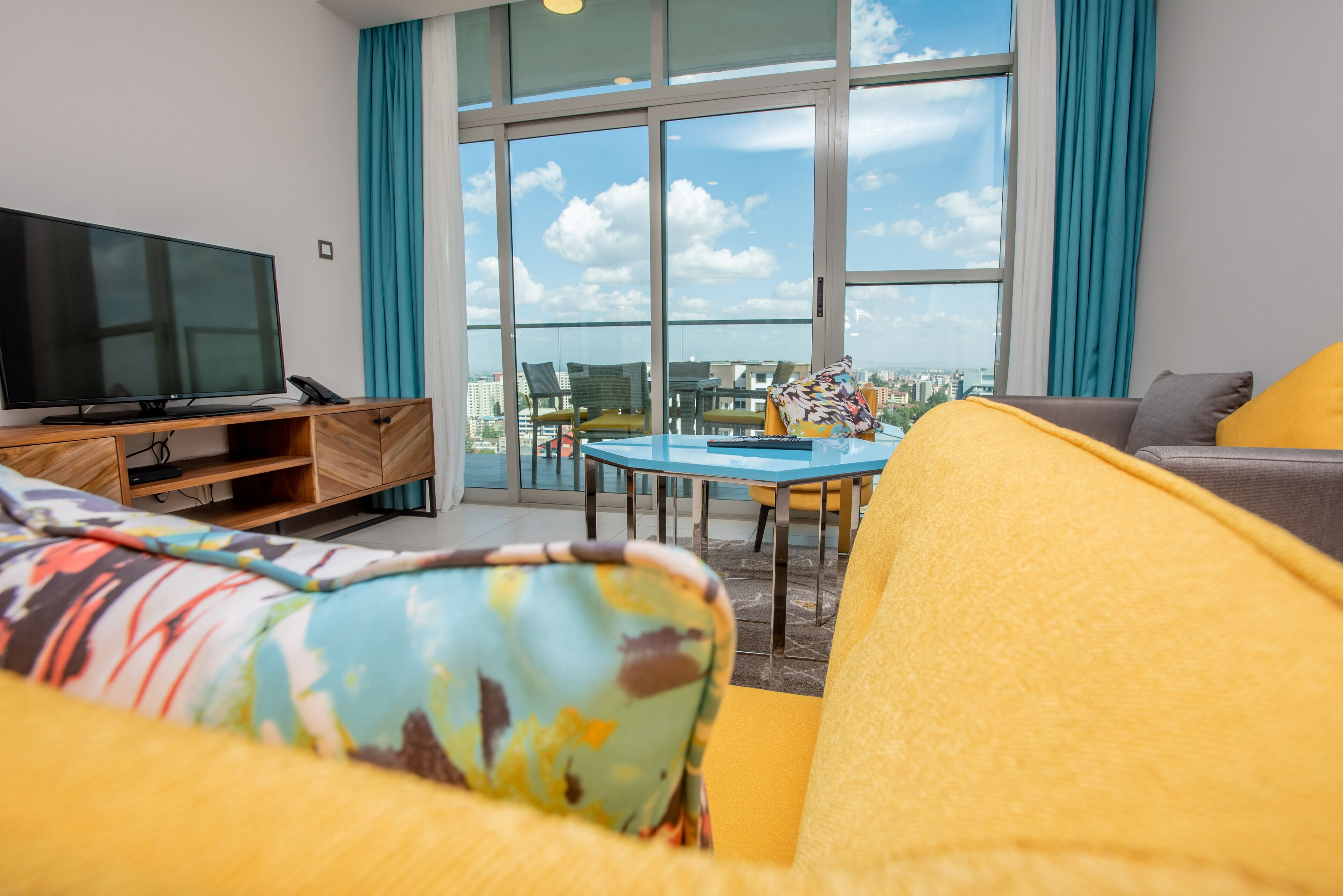 Ein modernes Wohnzimmer mit großem Fernseher, einer Couch, einem kleinen Tisch und einem Balkon mit Blick auf die Stadt.