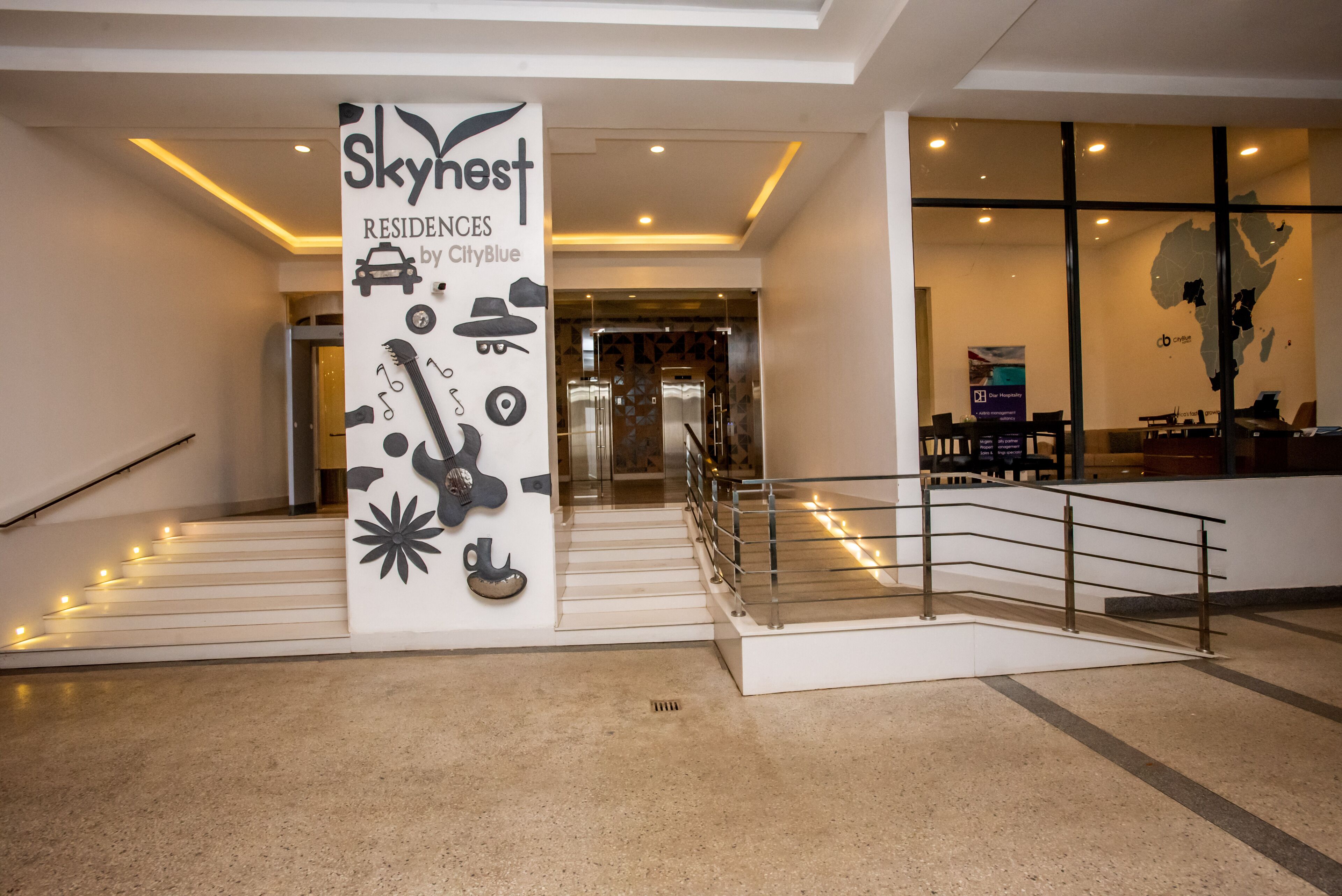 Um lobby de hotel moderno com uma coluna central ostentando o logotipo Skynest, uma escada com corrimão e uma área de recepção.