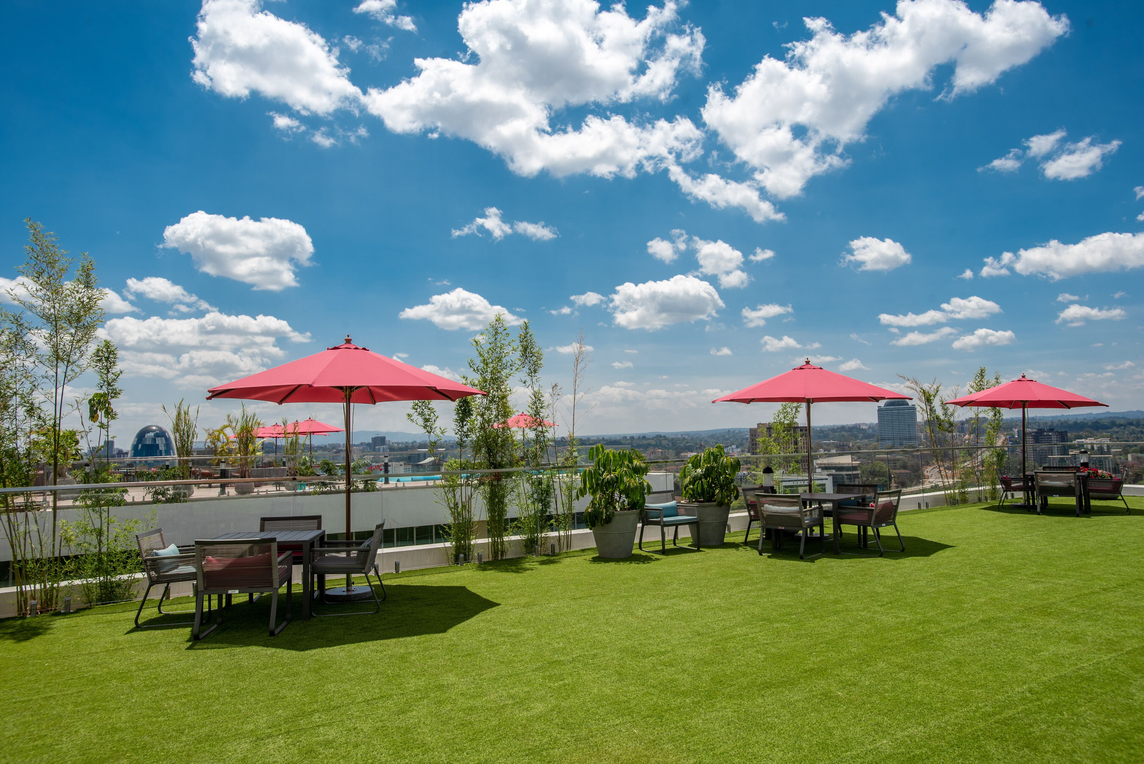 Patio esterno con ombrelloni rossi, prato verde e vista sullo skyline cittadino.