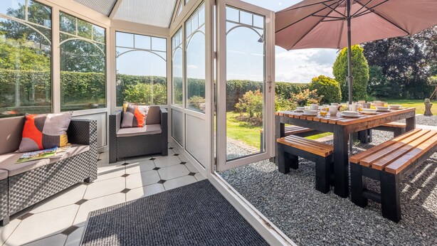 A conservatory with a glass door leading to a garden, furnished with a sofa and a table set for dining. - Routhorpe House (Driffield)