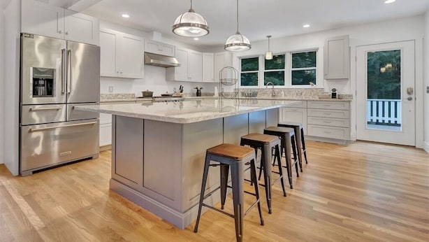 A modern kitchen with stainless steel appliances, a large island with bar stools, pendant lighting, and a view of the outdoors through large windows.