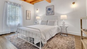 A bedroom with a white metal bed, two bedside tables with lamps, a large window with sheer curtains, and a wooden ceiling.