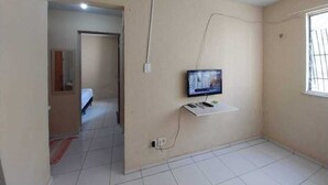 A room with a television mounted on the wall, a white shelf below it, and a window on the right. - APARTMENT NEAR SKYSURF CUMBUCO IN CAUCAIA (CAUCAIA)