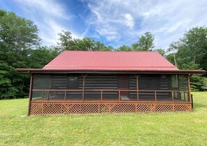 Situated in a serene grassy area, this cabin offers a spacious open porch with a lattice railing and screened area.