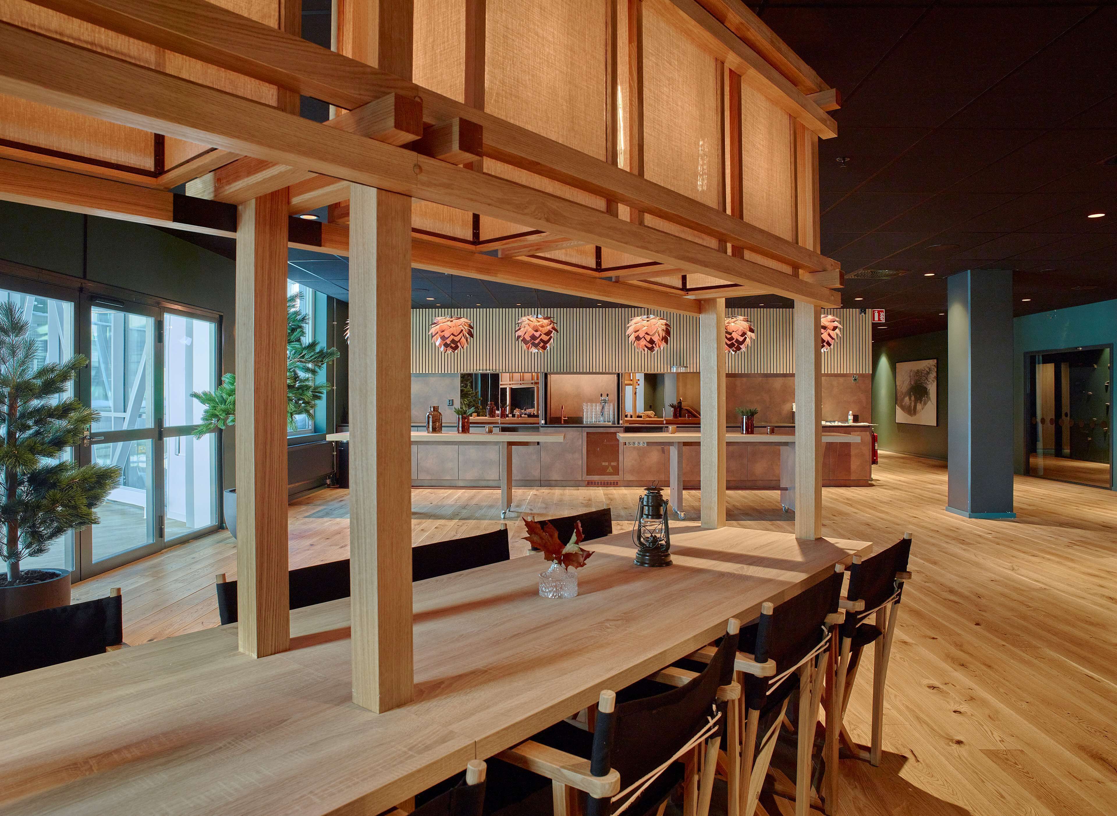A modern interior with wooden beams, a long table, and chairs. There is a kitchen area in the background with a large window.