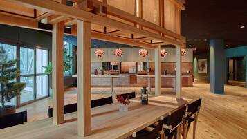 A modern interior with wooden beams, a long table, and chairs. There is a kitchen area in the background with a large window.