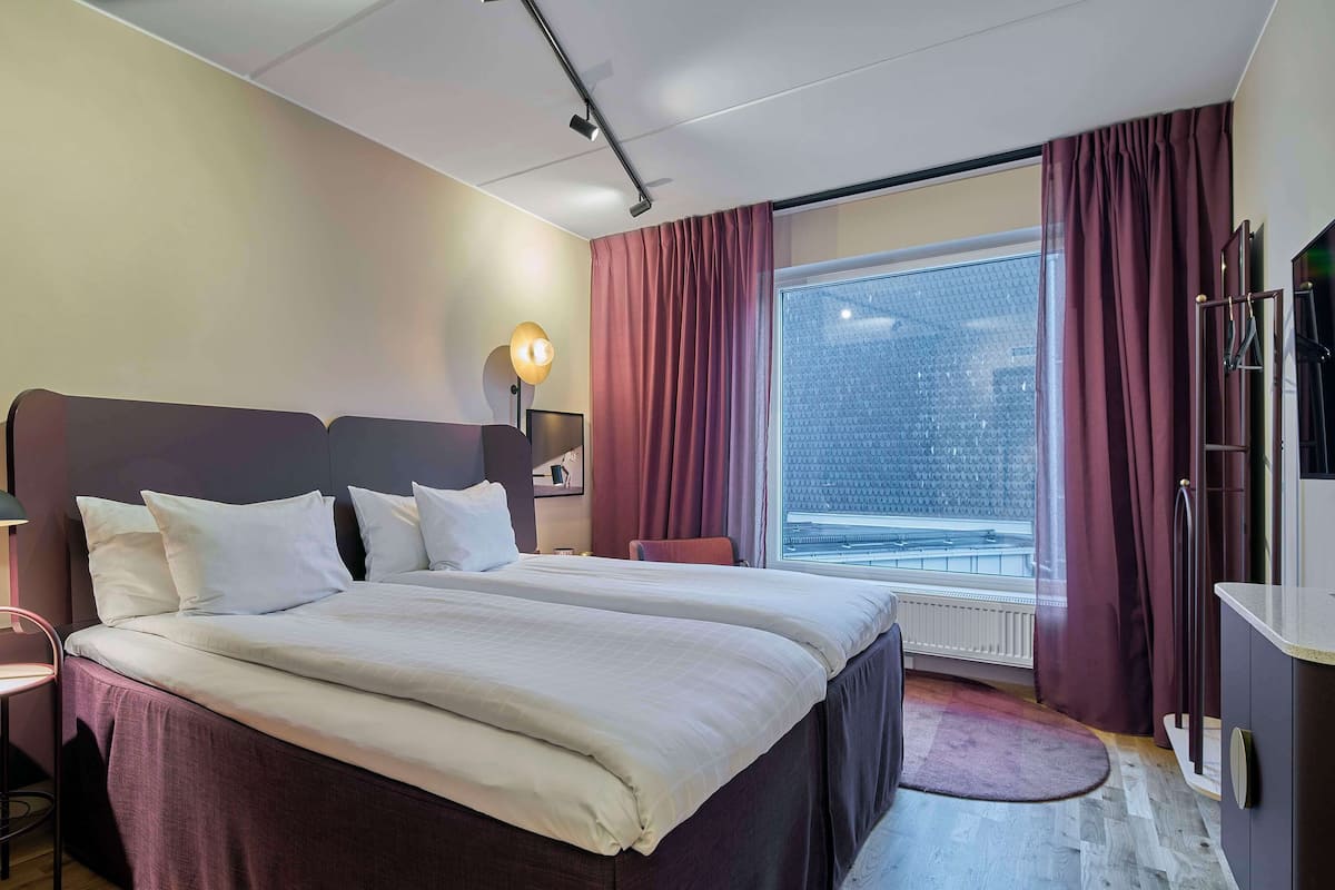 Une chambre d’hôtel avec un grand lit, un bureau, une chaise, une lampe de bureau, un téléviseur fixé au mur et une fenêtre avec des rideaux.