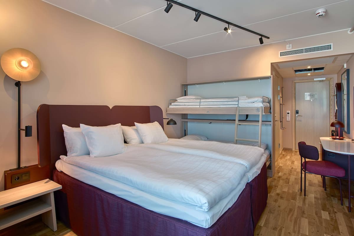 Une chambre d’hôtel avec un grand lit, un bureau avec une chaise et, au fond, un lit superposé.